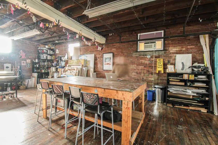 Studio space with tables, printing press, and artwork in the background. A blurry person stands at a laptop.