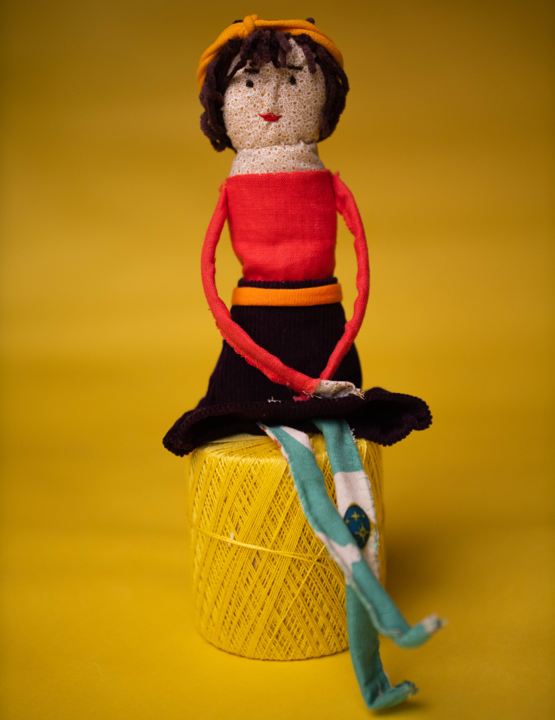 A doll with a red shirt, black skirt, and green legs, sits on a spool of yellow thread, on a yellow backdrop.