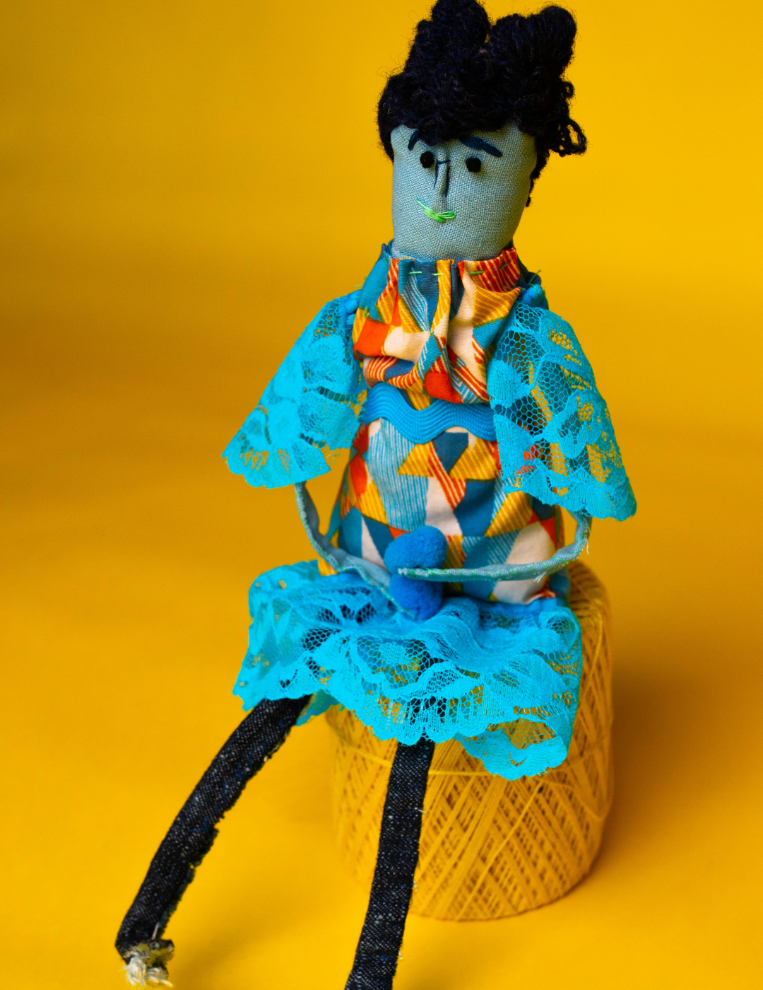 Person wearing a blue and orange dress, sits on a yellow spool of thread, on a yellow backdrop.