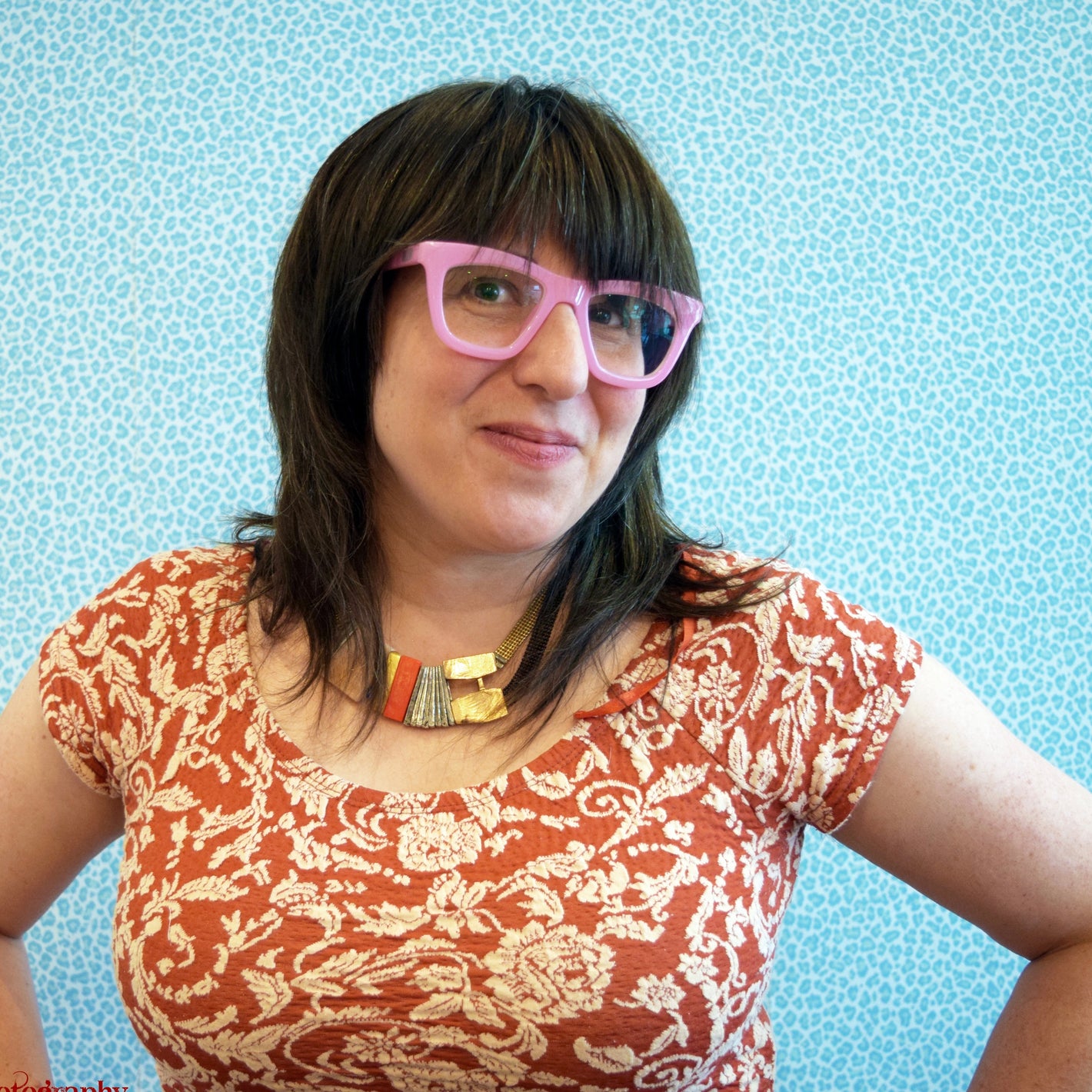 Person wearing a red patterned shirt and pink glasses, stands smiling in front of a blue patterned background.