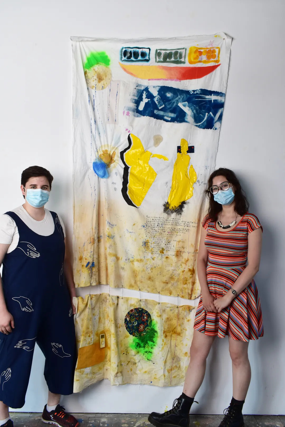 Two people wearing medical masks stand beside a piece of artwork hanging on the wall - two pieces of fabric hanging with abstract shapes and colours.