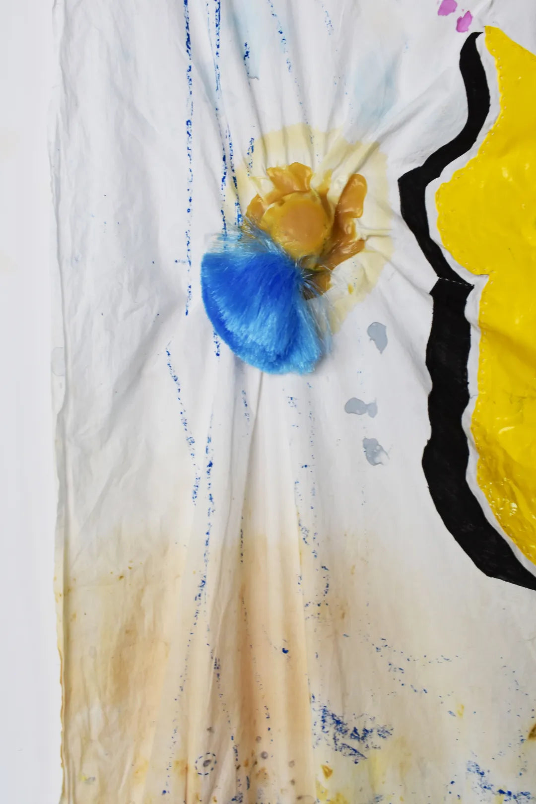 Close up of fabric with blue and yellow dyed ink splattered. A blue hairy tassle hangs off of a yellow sticky substance.