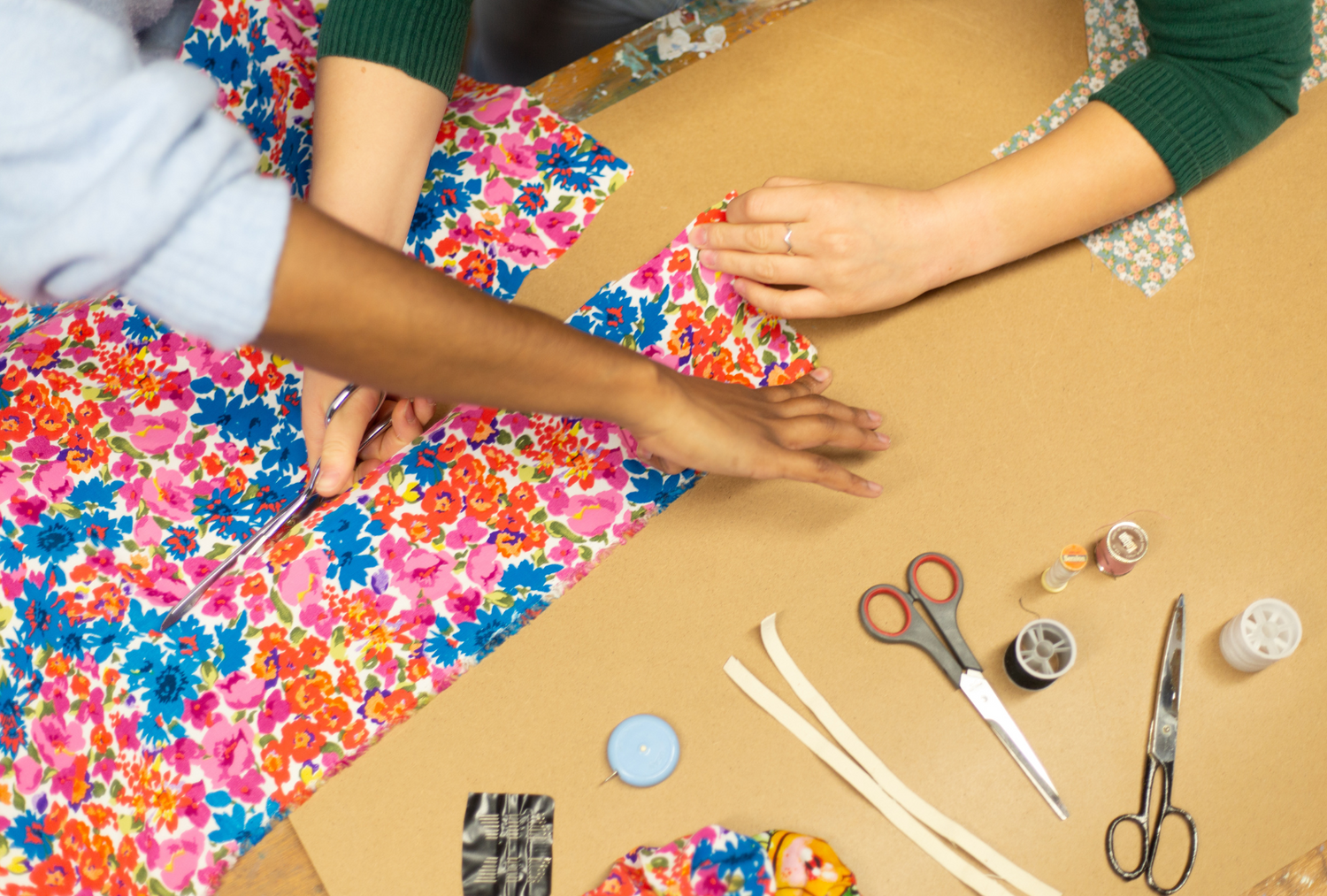 Two sets of hands hold floral print fabric while cutting it. Around the table sits scissors, thread, elastics, and other sewing materials.