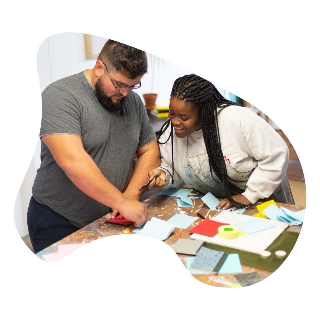 Two people work at a workshop table, one person smiles and points at what the other person is doing. Many pieces of paper and rolls of tape lie in front of them.