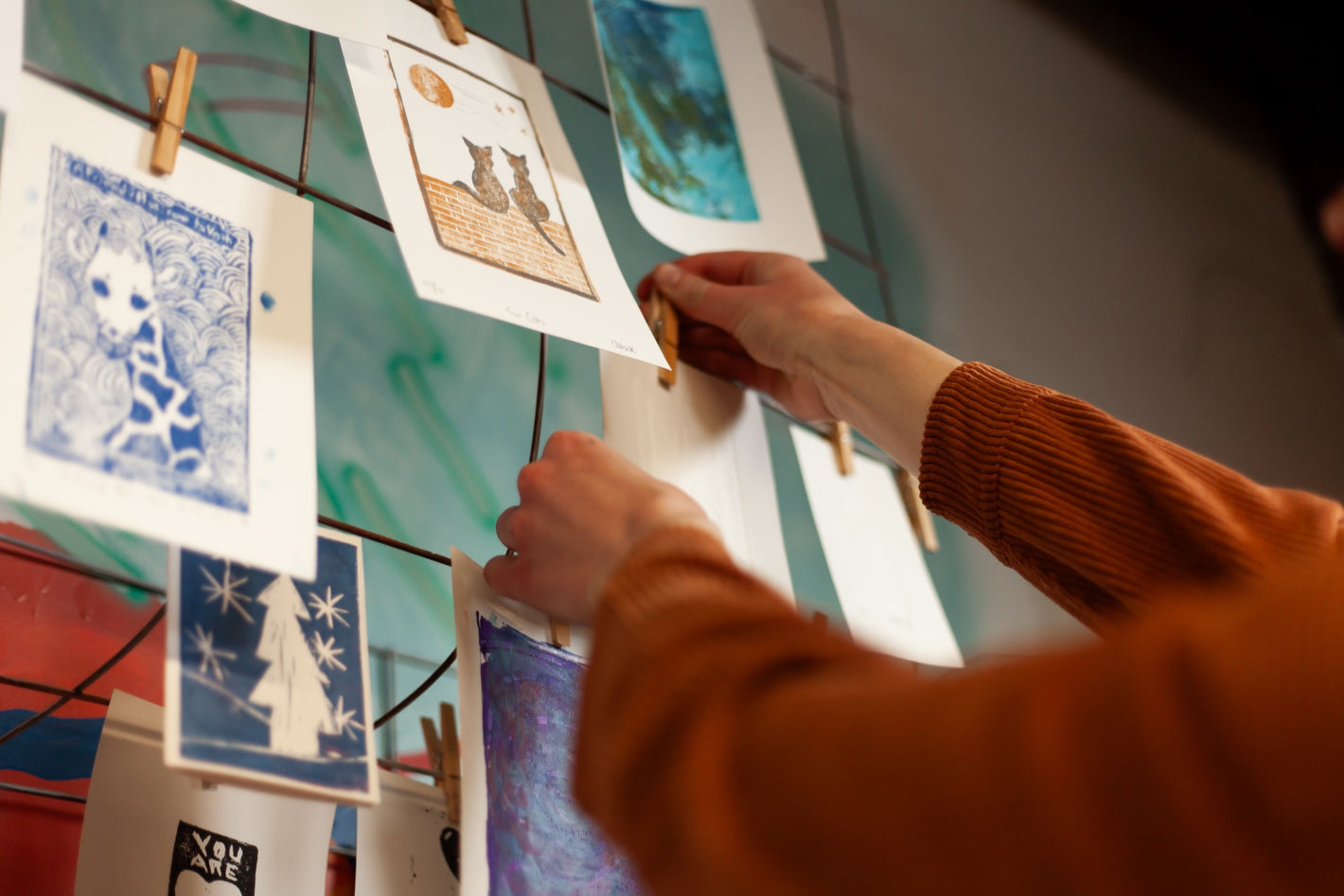 Close up on hands pinning a piece of paper up to a wire rack with many prints hanging using clothes pins.