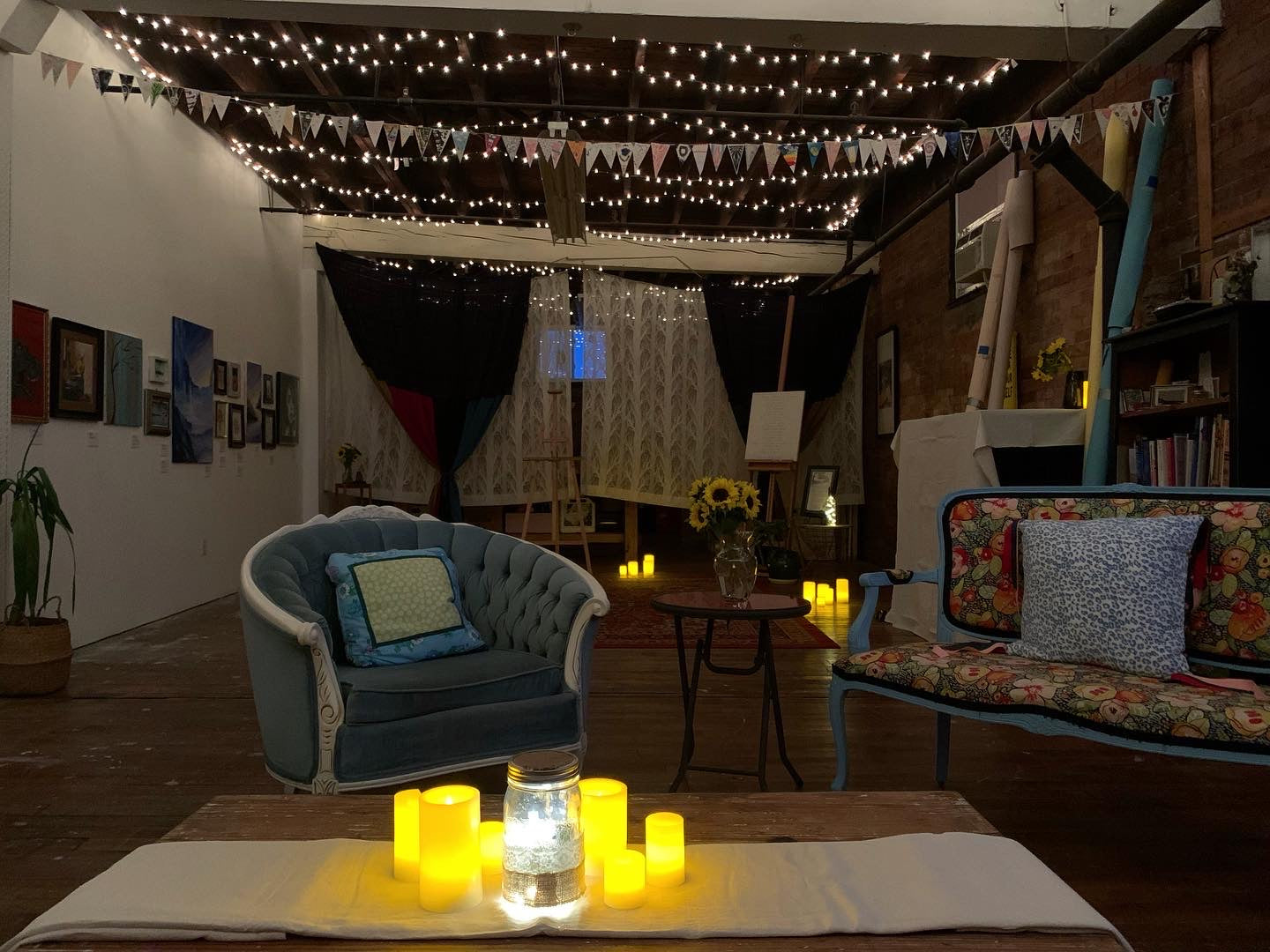 Otherwise studio dark, light up with candles and twinkly lights. Couches face each other, with sunflowers in the middle. Curtains hang in the background, and art hangs on the wall to the left.