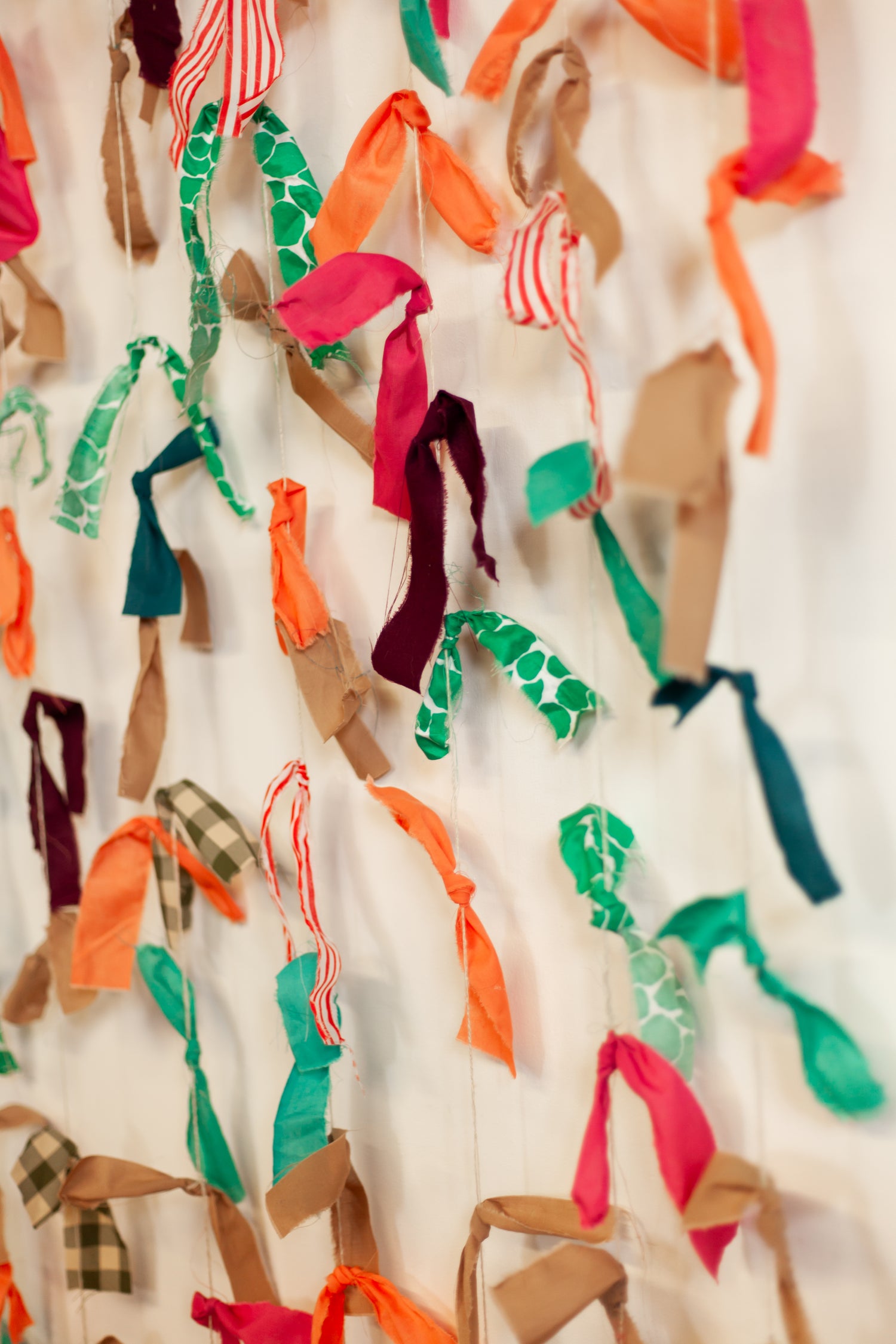 Colourful fabric in strips, tied to string hanging against a white wall.