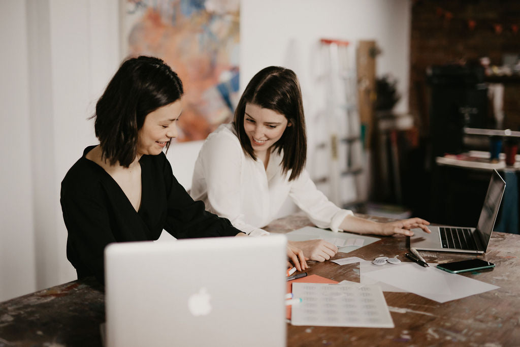 Two people sit at a paint-splattered table, smiling, with laptops in front of them and art on the wall behind them.