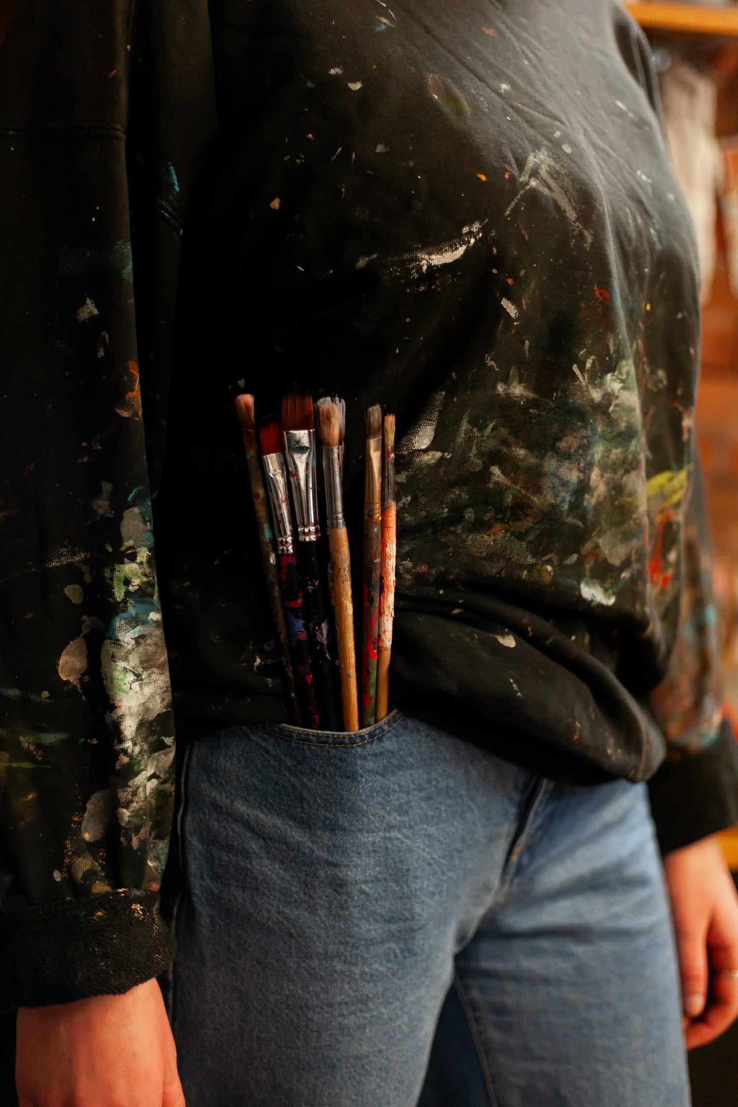 Closeup of paintbrushes sitting in jeans pocket on a person wearing jeans and a paint-covered sweater.