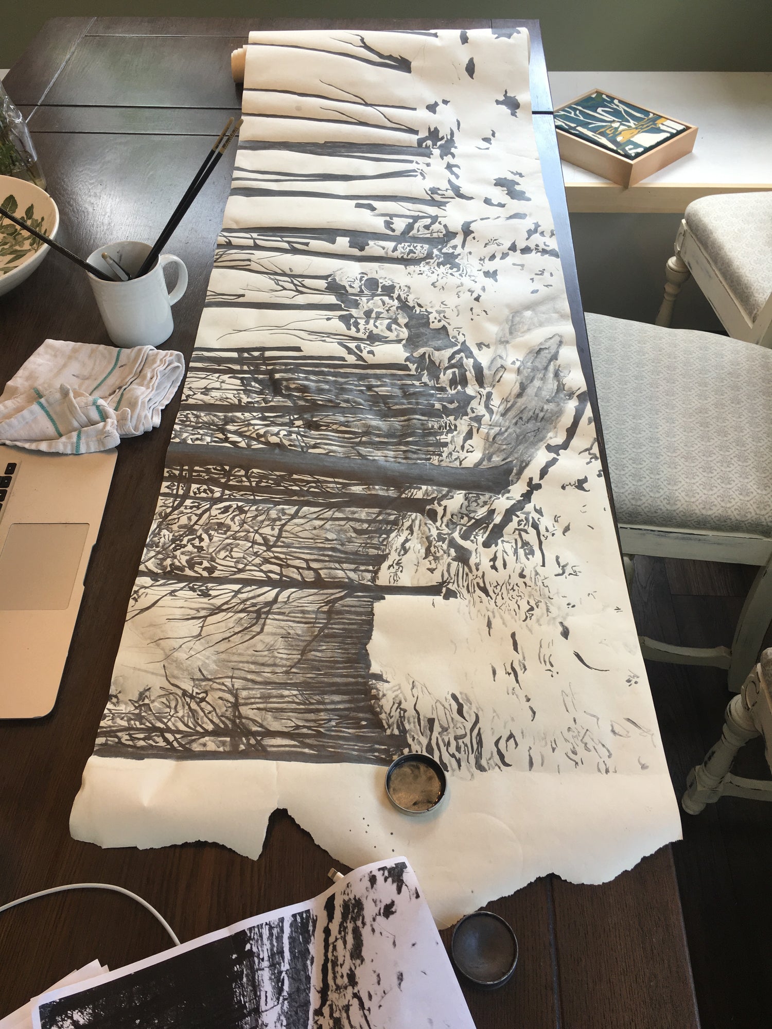 Long canvas rolled out on a table with black and white painted image of a landscape.