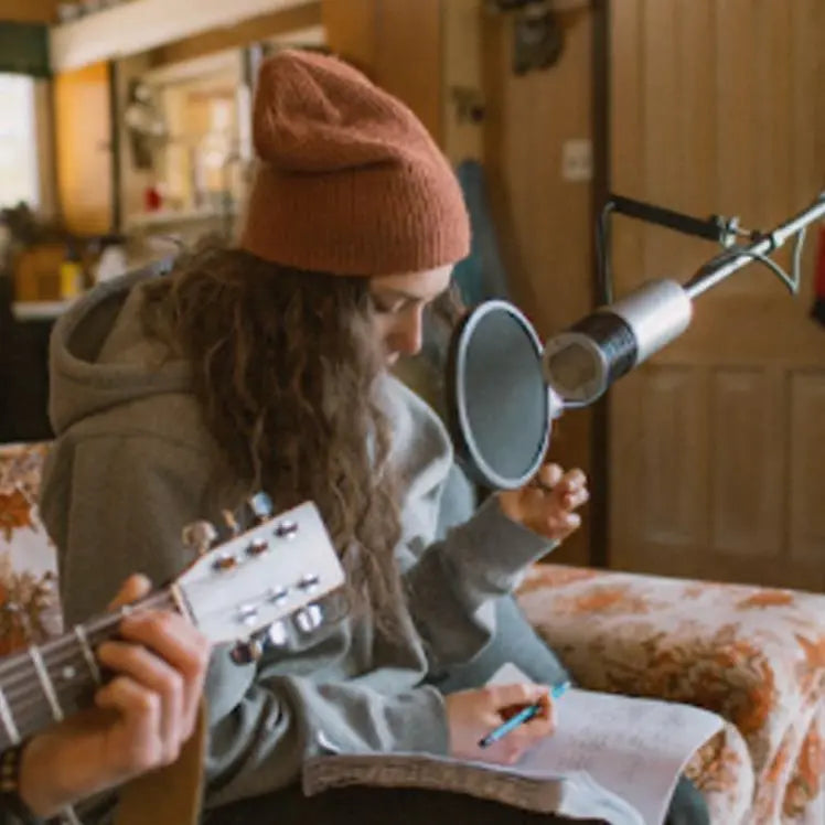 Person wearing a toque with long curly hair sits infront of a microphone, looking down at a notebook with a pencil in hand.