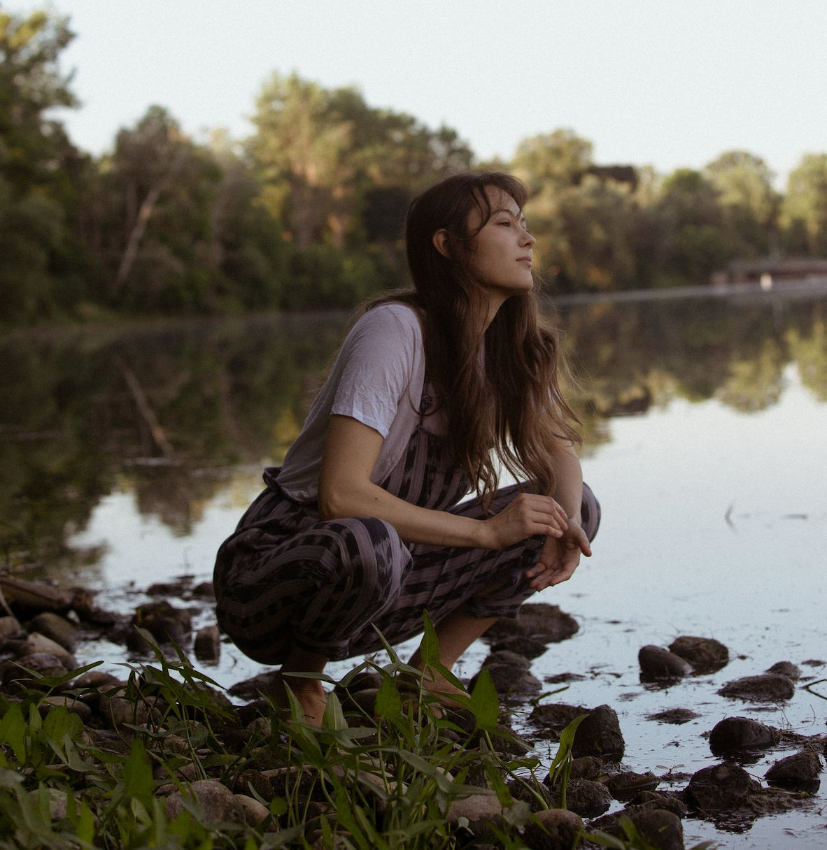 Person with long hair crouches by the water, looking out.