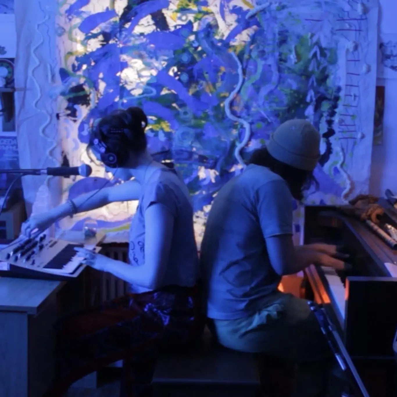 Two people sit with their backs against each other in a purple-lit room. One plays a piano and the other plays a synthesizer.