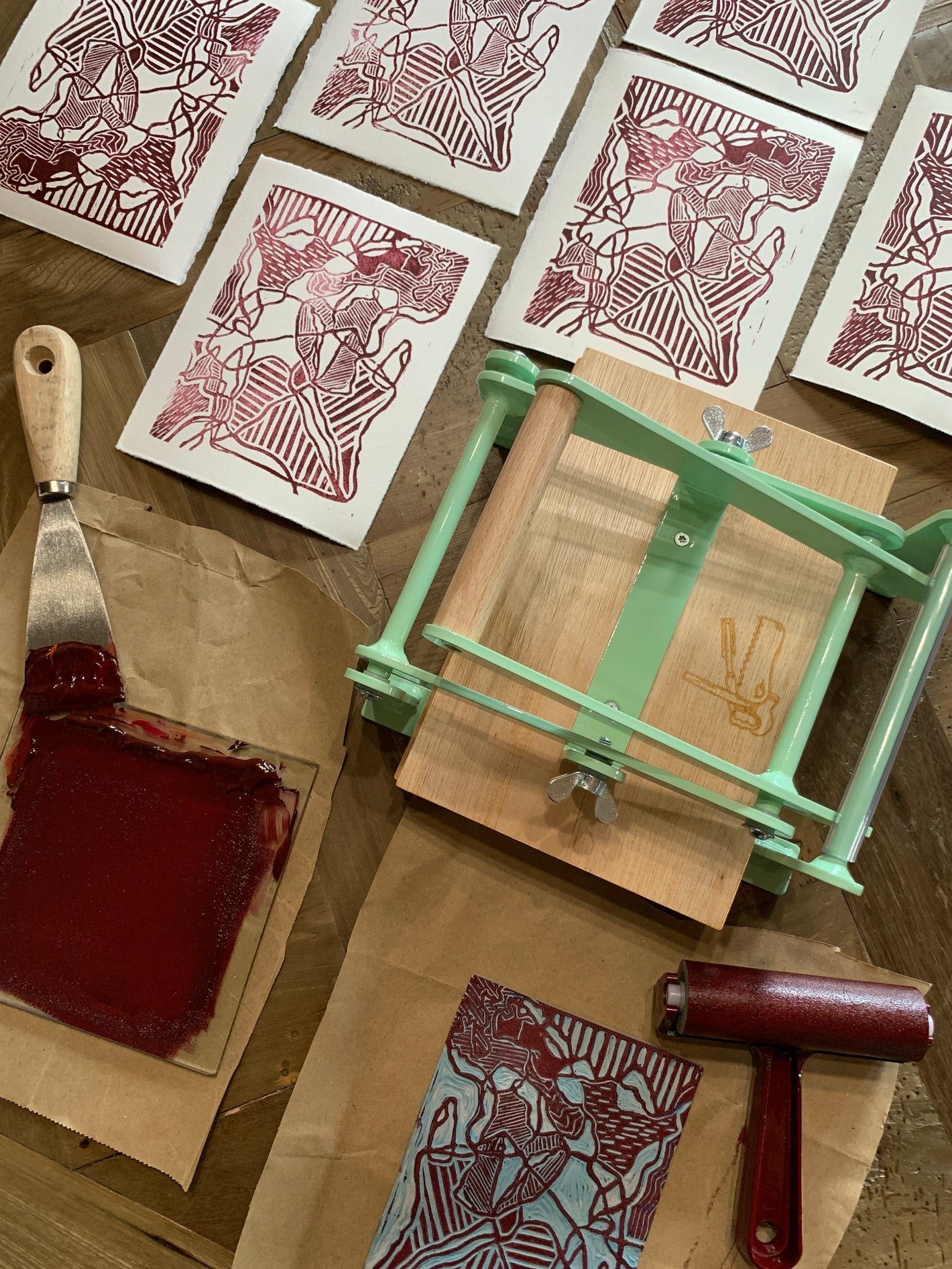 Birds eye view of linoblock prints lined up on a table, beside a small portable Woodzilla printing press  and dark red ink.