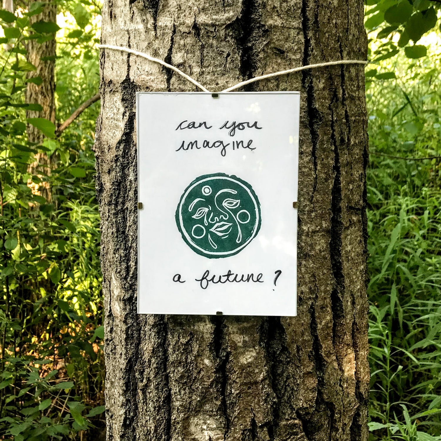 Printed image of a moon with a face on it, with text that reads "can you imagine a future?", tied around a tree.