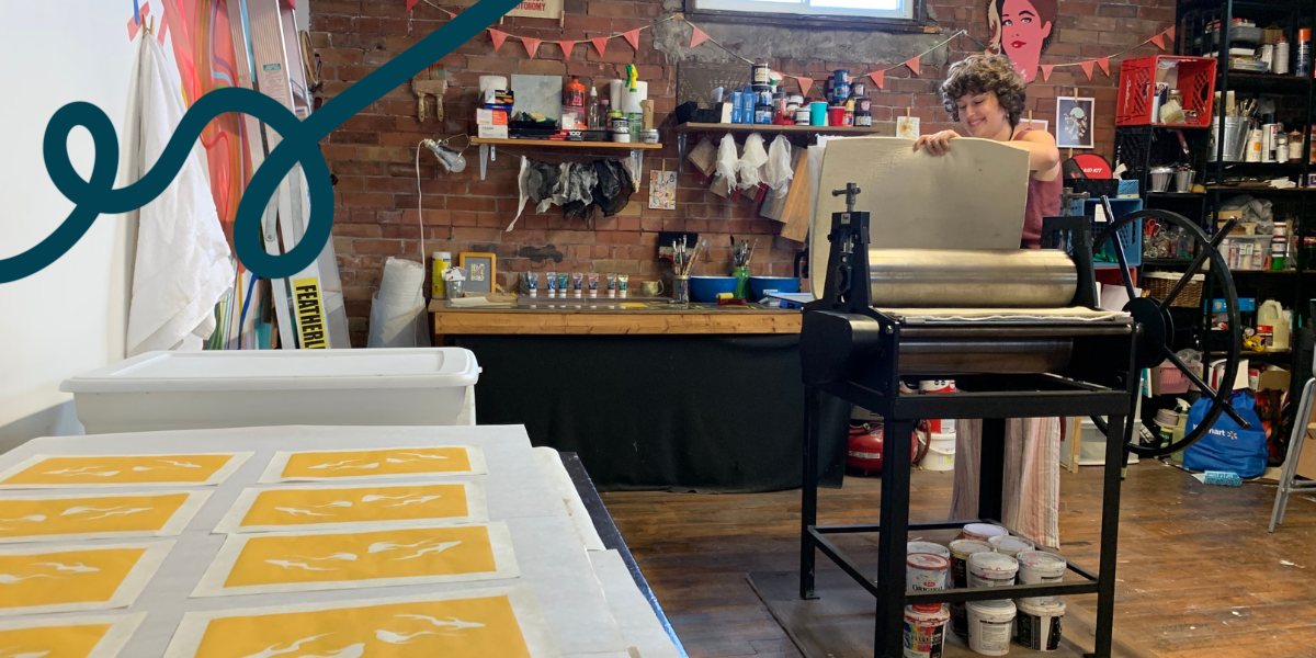 Person smiles while lifting up the blankets of a printing press. In the background are shelves and a table with printmaking inks and materials. In the foreground are yellow prints sitting on a table.