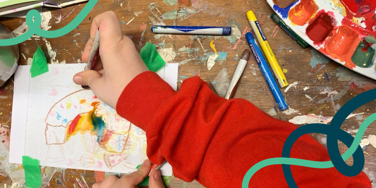 A small hand uses a water-soluble crayon on a piece of plexiglass, drawing half a donut. Navy blue and turquoise squiggly lines decorate the image.