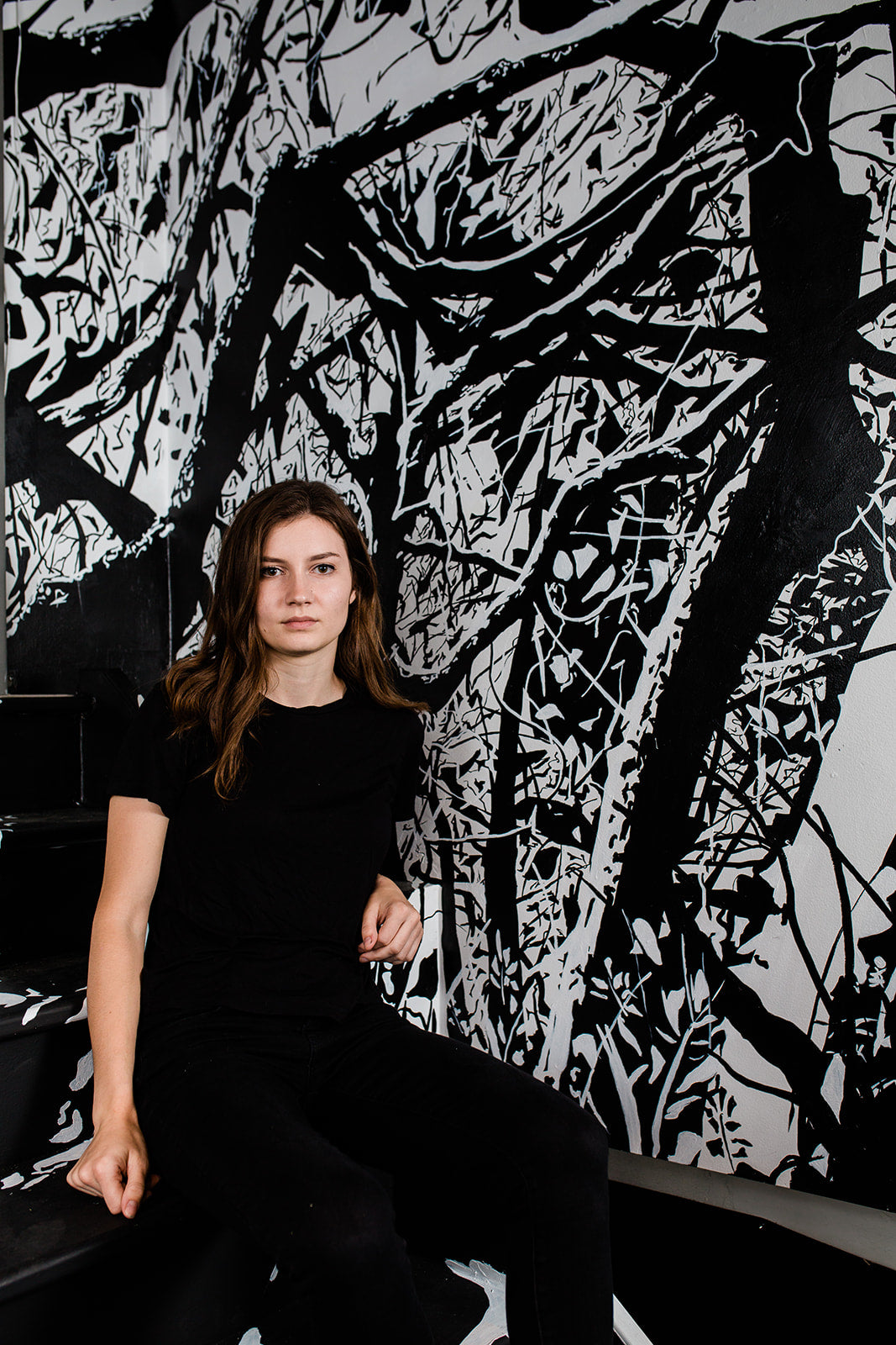 Image of a person in all black staring seriously at a camera, in front of a wall with black and white painted landscape imagery.