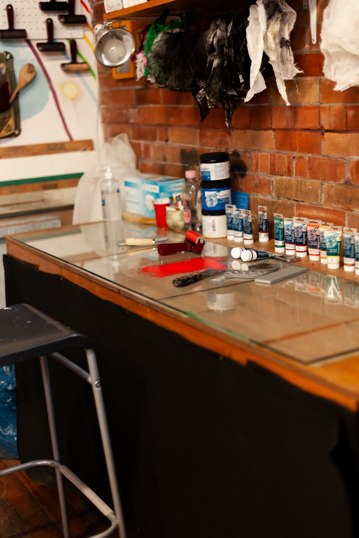 View of a printmaking table with red ink spread out, and tools beside the ink including a brayer and palette knife. Lined up against the brick wall are many inks and rags.