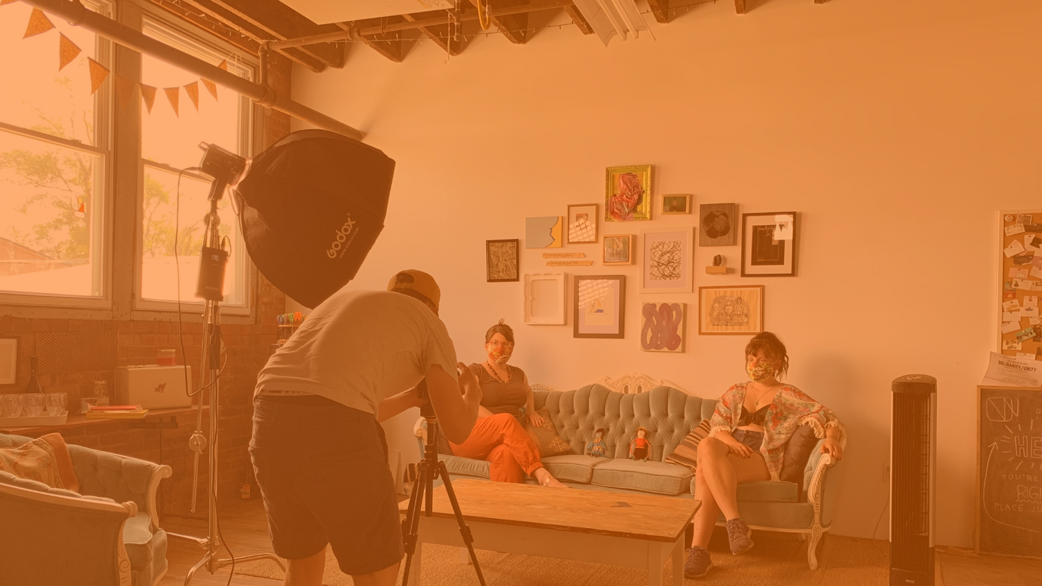 Orange-overlayed image of two people wearing medical masks, sitting on a couch separated from each other. A person in front of them is taking a photo.