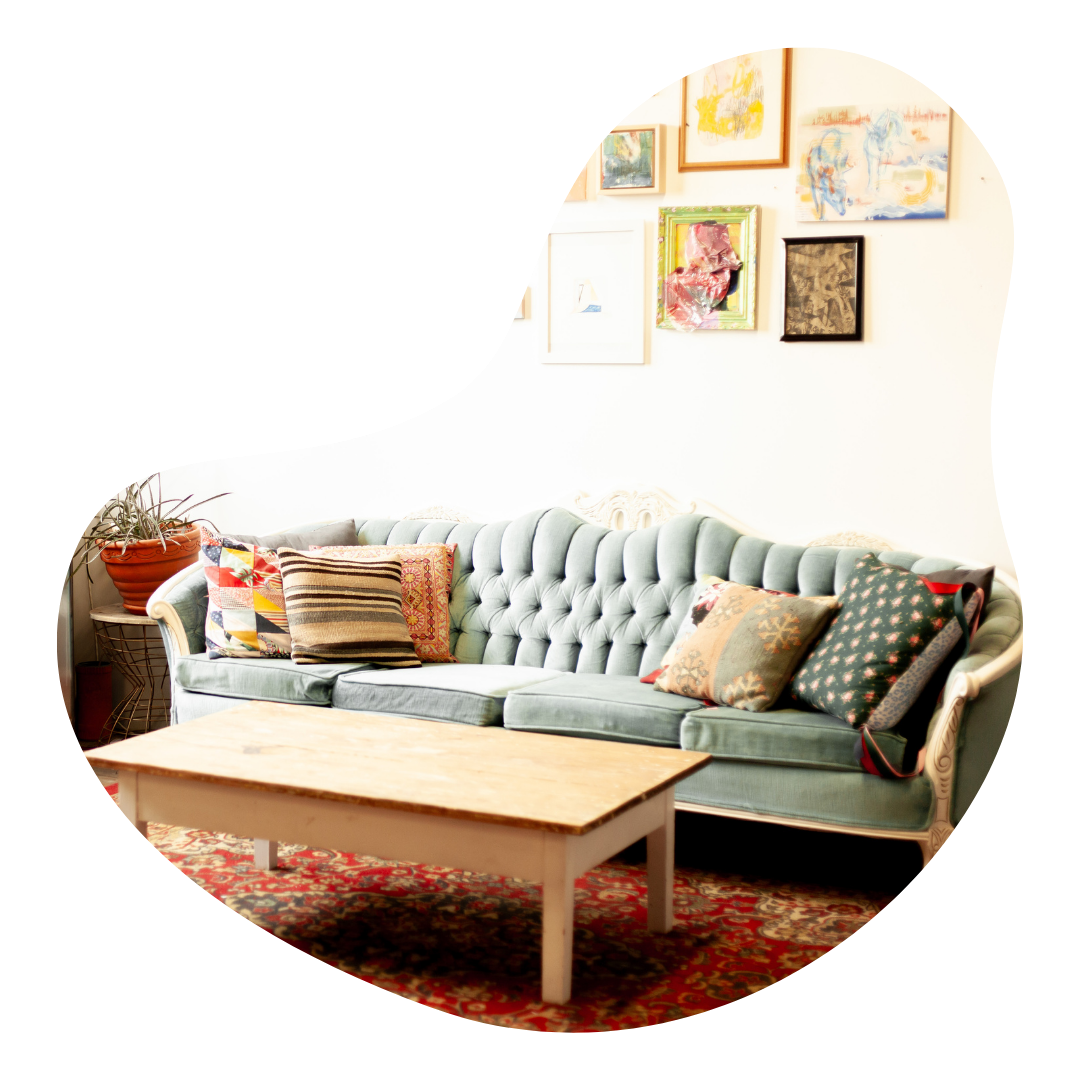 A turquoise vintage couch with pillows, with a wooden coffee table in front. On the wall behind is a salon style wall with many pieces of art hanging.