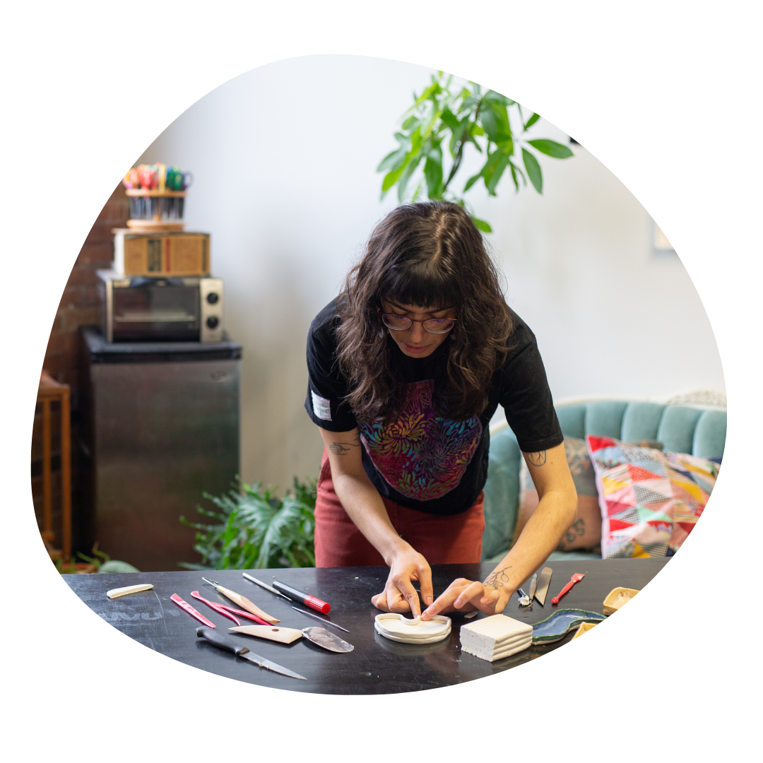 Person hand builds a ceramic tray at a black table. On the table are various clay building tools. In the background is a green plant and art materials.