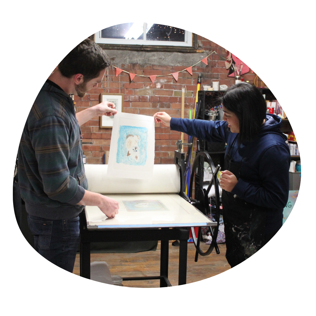 Two people lift a monoprint of a cat off of the printing press in front of a brick wall.