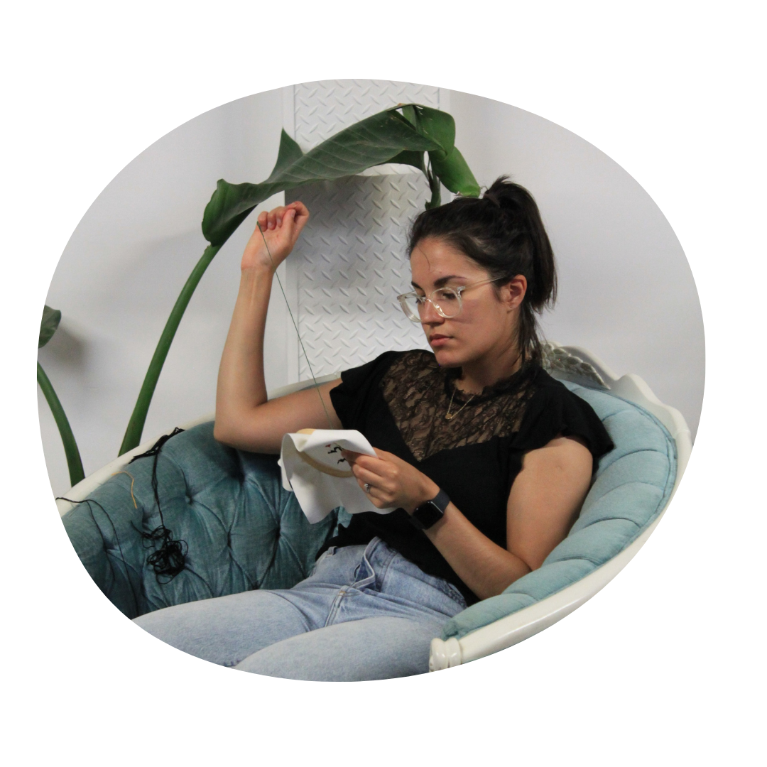 Person with glasses and hair in a ponytail sits in a blue vintage chair, pulling thread through an embroidery hoop.