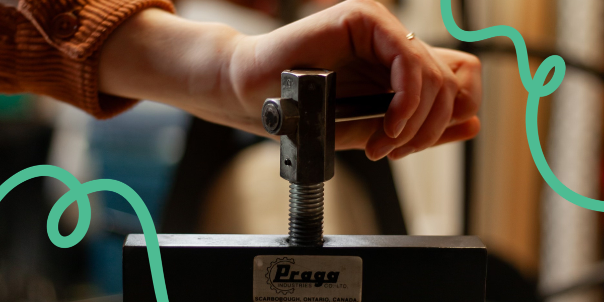 Close-up of a hand tightening the pressure on a Praga intaglio press. On either side of the hand, squiggly turquoise lines decorate the image.