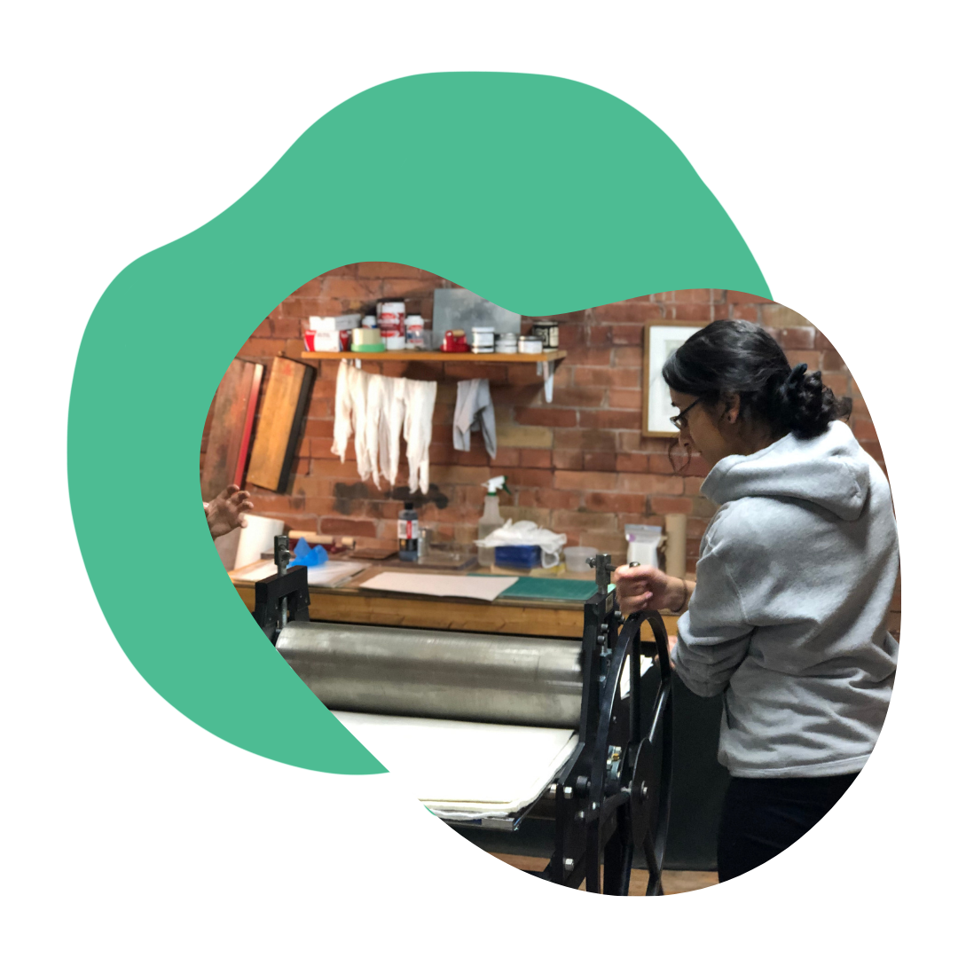 Person moving the wheel of a printing press with brick wall behind them and printmaking materials. Turquoise blob shape behind the image as an accent.