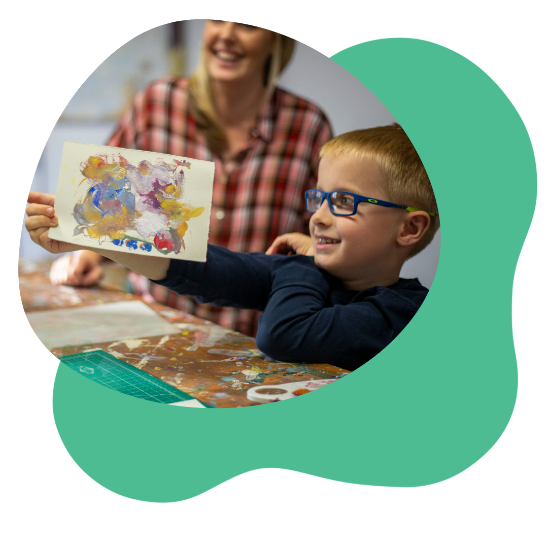 Young kid with glasses smiles and looks at a print they made. In the background, an adult smiles and is blurred out.