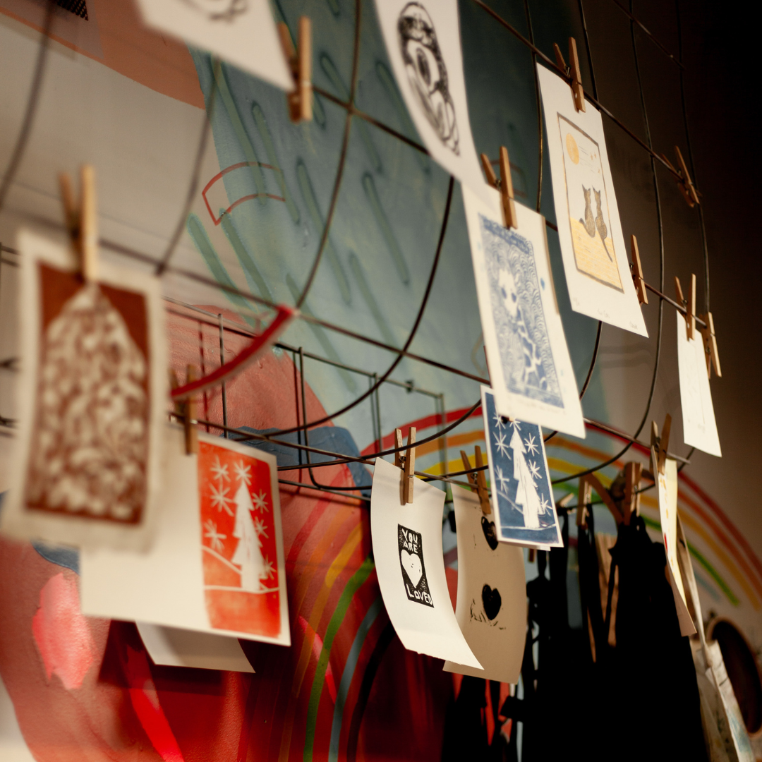 Colourful prints hang with clothespins on a metal frame on a colourful wall with a rainbow. Aprons hang on the metal frame on the right.