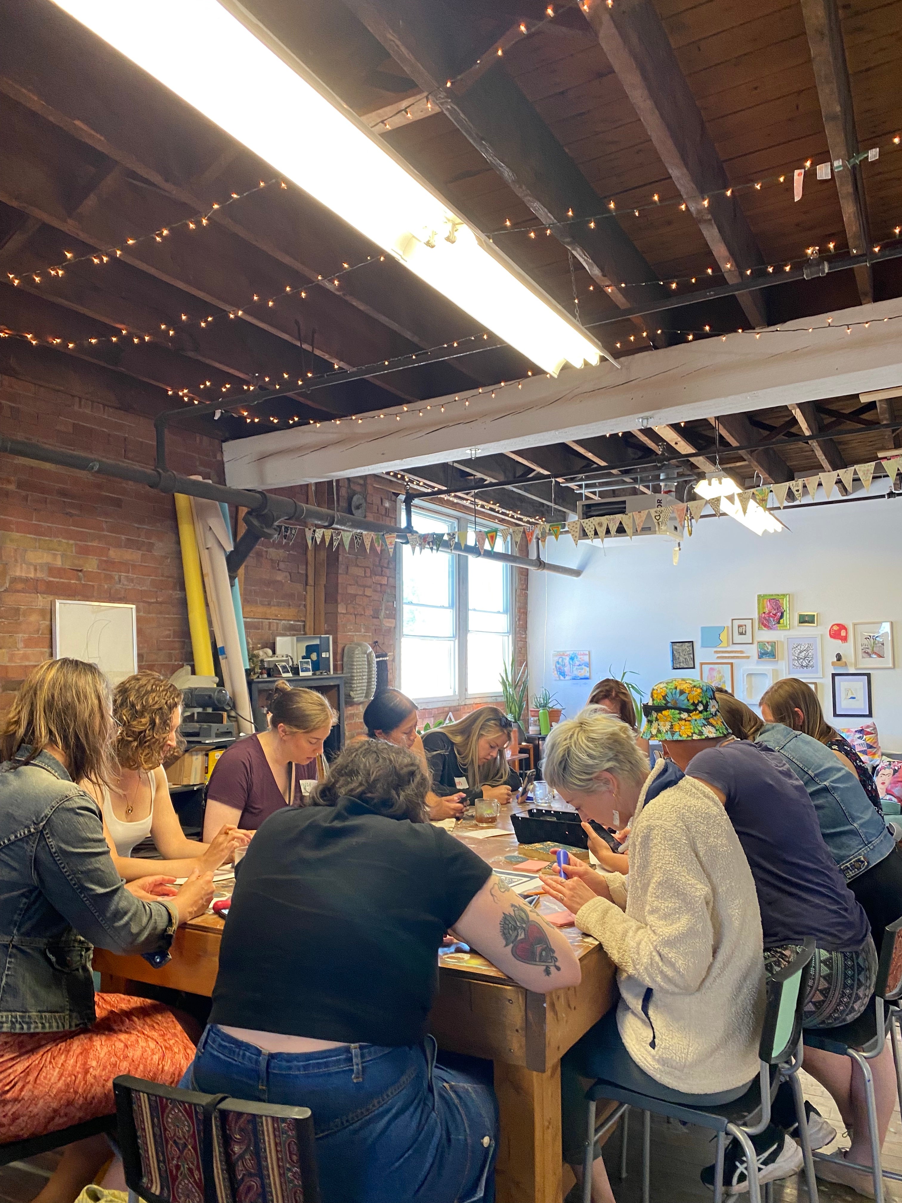 A group of people sit around a table working an linocut prints. Twinkly lights light up the ceiling.