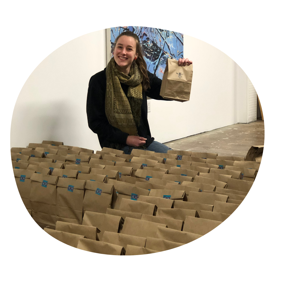 Person smiles while holding up a paper brown bag. In front of them, several brown bags sit on the ground.