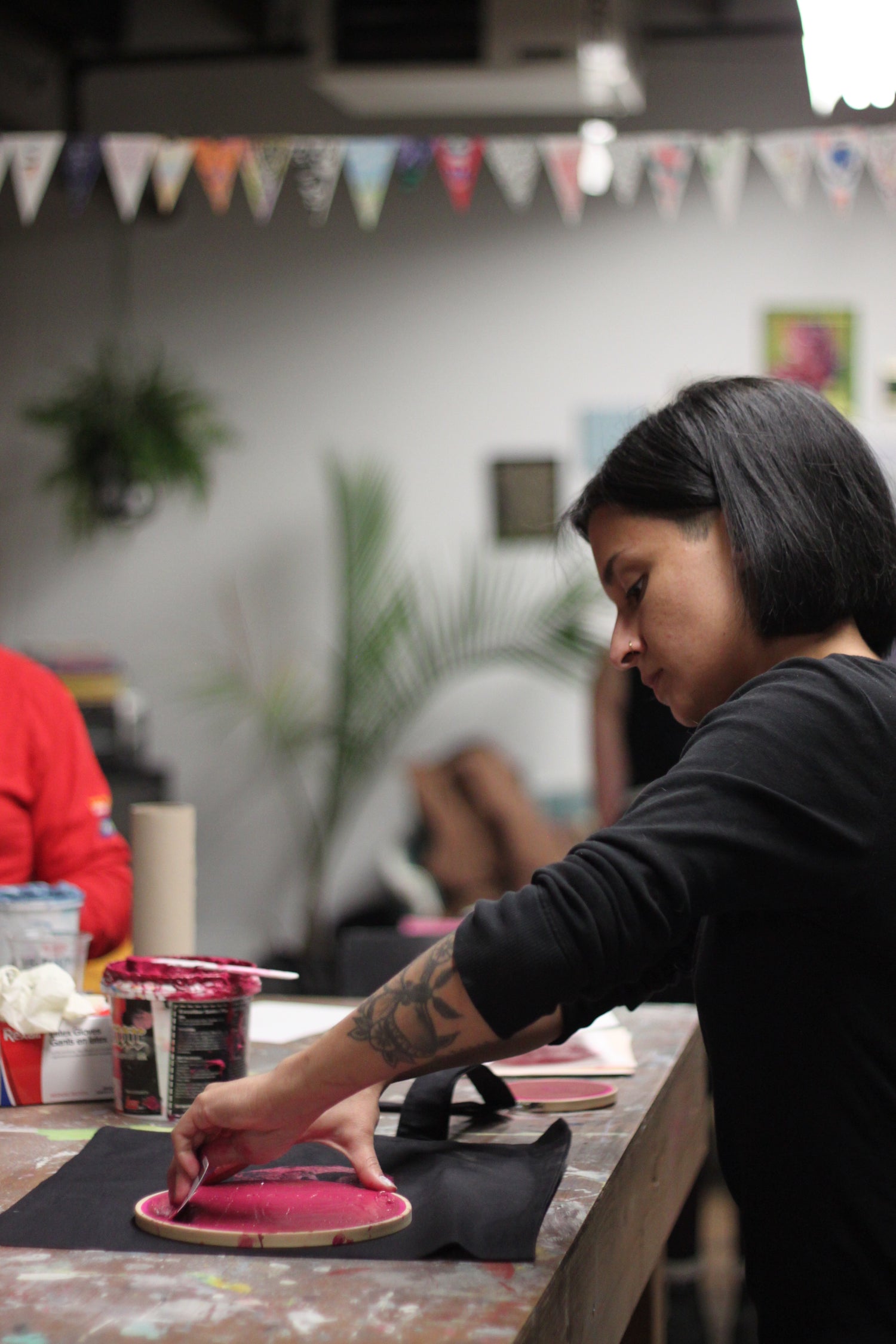 Person pulls ink through a DIY round screenprinting frame. In the background is a bunting banner blurred out.